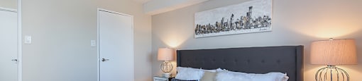 Primary bedroom, looking towards closet, with a grey upholstered bed frame and white linens at Lorneville Apartments in Cornwall, ON
