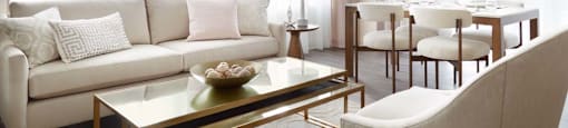 A modern living room with a white sofa, a glass coffee table, and a dining area in the background.