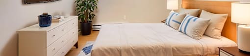A bedroom with a bed, nightstand, and a window with a view of the outside at Moira Apartments in Belleville, ON