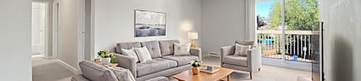 A living room with a grey couch, a grey ottoman, a grey sofa, a grey coffee table, a grey rug, a grey wall, a white door, a white wall, a white ceiling, a white floor at Moira Apartments in Belleville, ON