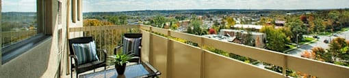 Edgehill Apartments in Barrie, ON balcony view overlooking lush trees and homes. The weather is clear with bright blue skies and two chairs are sat next to each other with a glass table in front.