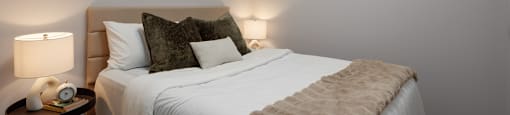 A bedroom with a bed, pillows, a blanket, a nightstand with a lamp, and a painting on the wall at The Ambrosi in Kelowna, BC.