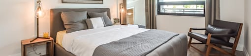 Bedroom with a large, grey upholstered bed and a window in a 1 bed 1 bath variation B in building A at Trio on Belmont in Kitchener, ON