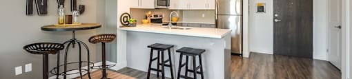 Two bars at a breakfast bar in an open-concept kitchen, with a dinette to the left, in a 1 bed 1 bath variation B in building A at Trio on Belmont in Kitchener, ON