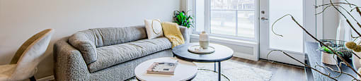 A living room with a grey couch, two round coffee tables, large window and door to a patio in a 2 bedroom 2 bathroom variation C layout at Vale Station Apartments in Waterloo, Ontario