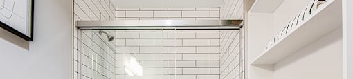 Main bathroom with a white tiled shower with a glass door, white toilet and shelving in a 2 bedroom 2 bathroom variation C layout at Vale Station Apartments in Waterloo, Ontario.