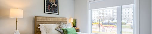 Second bedroom with a large bed, a nightstand with a lamp, and a picture on the wall next to a large widow with blinds in a 2 bedroom 2 bathroom variation C layout at Vale Station Apartments in Waterloo, Ontario