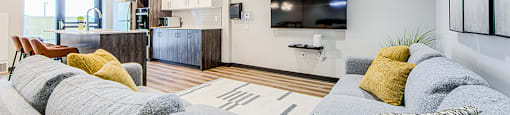 Large grey sectional couch and flat screen TV with a kitchen in the background in the social room at Vale Station Apartments in Waterloo, Ontario
