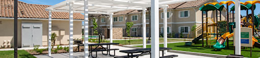 A white pergola with picnic tables is in the foreground of a playground.
