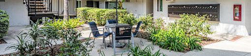 A patio with a table and chairs is surrounded by plants and a building with balconies is in the background at The Marquee Apartments, California 91605