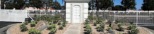 A small white building with a black roof is surrounded by a white fence and palm trees.