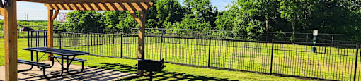 Wooden pergola with a picnic table is situated in a grassy area with a fence in the background.