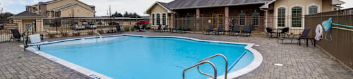 Large outdoor swimming pool surrounded by a brick patio and a house with a black roof.