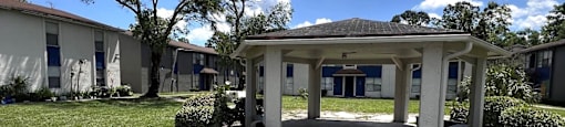 A gazebo is in the middle of a grassy area with apartment buildings in the background.