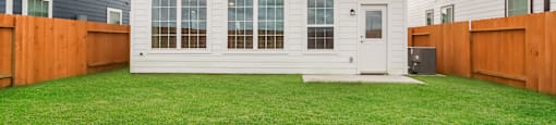 A white house with a brown door and windows is surrounded by a green lawn and wooden fences.