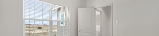 A room with a carpeted floor, a window with blinds, and a door leading to another room.