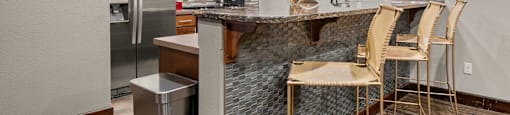 A kitchen with a refrigerator, microwave, brown cabinets, and a bar with chairs.