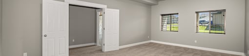 A room with a carpeted floor, windows with blinds, and a doorway leading to another room.