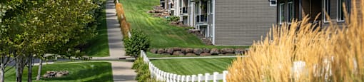 A sidewalk, a white fence, and trees behind a row of houses with backyards.
