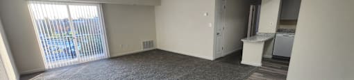 A spacious living room with a grey carpet and a kitchen area in the background.