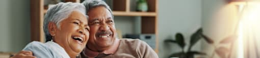 An older couple is sitting on a couch, smiling and laughing together.