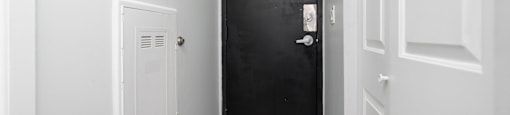 A black door with a silver handle is in the middle of a hallway with white walls and doors.
