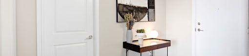A room with a white door, a table with a vase and a lamp, and a picture on the wall.