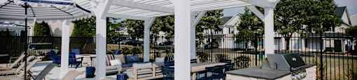 A white pergola is over a patio with chairs and a grill.