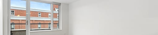 Empty room with wooden floors and a window overlooking a brick building.