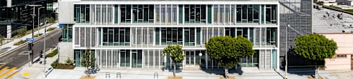 A modern building with a glass facade is situated on a street corner with trees and other buildings in the background.