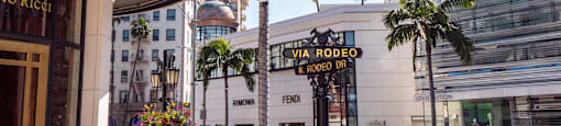 A palm tree stands in front of a white building with a sign that reads "VIA RODEO & RODEO DR.".