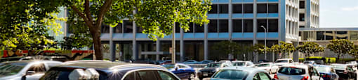 A tall building with many windows is surrounded by trees and cars.