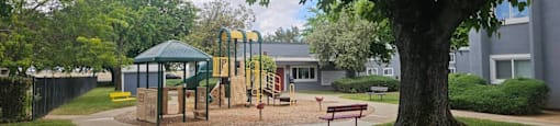 A playground with a green canopy and yellow slide is surrounded by a fence and a large tree.