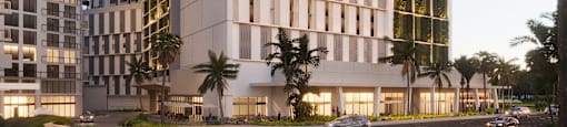 A modern building with a white facade and balconies is surrounded by palm trees and other greenery.