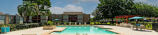 Gated community pool with lounge chairs, seating under umbrellas, and grilling station under pergola