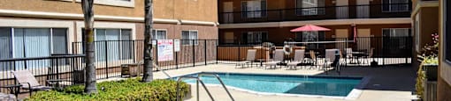 View of gated community pool and spa with lounge chairs and seating under an umbrella. Exterior of building in background