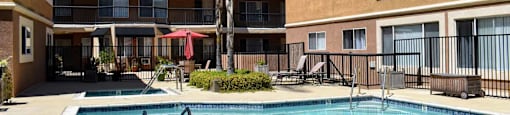 View of gated community pool with lounge chairs and seating area under an umbrella with exterior buildings andtropical landscaping