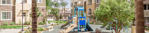A playground with a blue slide is surrounded by palm trees and apartment buildings.