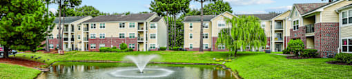 A pond with a fountain in the middle of a grassy area in front of apartment buildings.