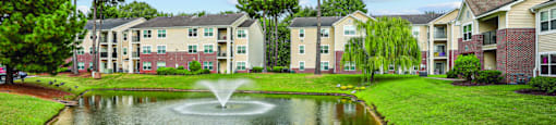 A pond with a fountain in the middle of a grassy area in front of apartment buildings.