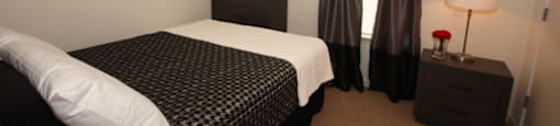 The furnished bedroom is carpeted with a queen bed, dresser and nightstand. The bed has a houndstooth comforter with white throw blanket, and a white pillow. There are black curtains hanging on the window and framed artwork on the wall.
