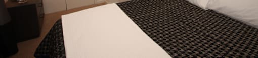The furnished bedroom is carpeted with a queen bedr and nightstand. The bed has a houndstooth comforter with white throw blanket, and a white pillow.