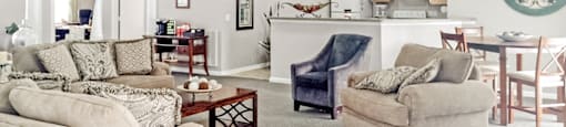 The resident clubhouse features ample seating including a couch, loveseat, and two chairs There is a white fireplace with a television mounted above it.. The room features gray carpet with beige walls that are set against a white textured ceiling with recessed lighting