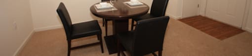 The furnished dining room has hardwood style flooring by the entry door, and carpet throughout the rest. There is a round wooden table with four black chairs positioned around it. There is a place setting for two on top of the table.