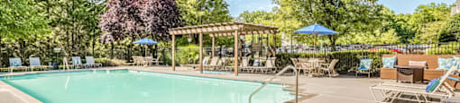 The community pool is rectangular in shape. The large sundeck area is enclosed with a black fence and features beige pool furniture including chaise lounge chairs, regular chairs, round tables, and blue umbrellas as well as a large wood-stained pergola. The exterior of the pool area is surrounded by bushes and mature trees. The community parking area is in the distance.