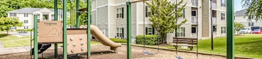 The community playground features a yellow, tan, and green play structure with a slide, a climbing wall, and two seated swings. The playground is surrounded by mulch, has a bench for seating, and sits adjacent to an apartment building in the background.