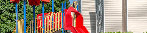 Red Yellow and Blue Playground with Red Seesaw with Building Exterior and Treeline in the Background