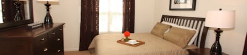The furnished mater bedroom is carpeted throughout with a large picture window on one of the walls. Featured here is a queen size bed with tan comforter and coordinating pillows, espresso colored dresser and nightstand, and a single lamp on top of each.