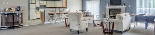 The furnished clubhouse is a large open room with a coffee bar, a full kitchen with breakfast bar, and living area with a fireplace and two sliding glass doors on either side of the fireplace. It is furnished with three bar stools, two couches and an oversized chair, two smaller tables with chairs, and a coffee table.