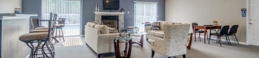 The furnished clubhouse is a large open room with a coffee bar, a full kitchen with breakfast bar, and living area with a fireplace and two sliding glass doors on either side of the fireplace. It is furnished with three bar stools, two couches and an oversized chair, two smaller tables with chairs, and a coffee table.